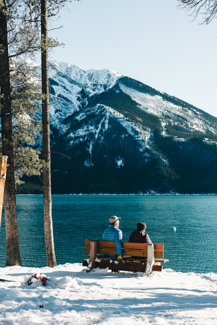 Two people enjoy a serene view of a snowy mountain and lake in Banff, Alberta, Canada.